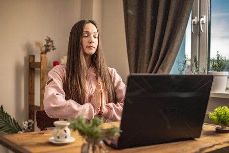 Woman is sitting at a table in front of a laptop, meditating and relaxing while listening to an instructor online or a video lecture. Concept of a remote therapy and meditation session.の写真素材