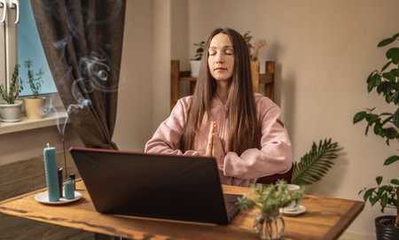 Woman is sitting at a table in front of a laptop, meditating and relaxing while listening to an instructor online or a video lecture. Concept of a remote therapy and meditation session.の写真素材