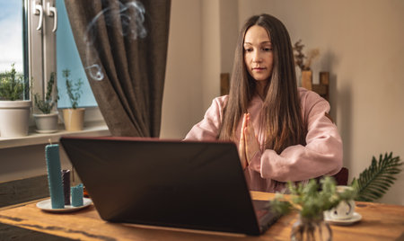 Woman is sitting at a table in front of a laptop, meditating and relaxing while listening to an instructor online or a video lecture. Concept of a remote therapy and meditation session.の写真素材