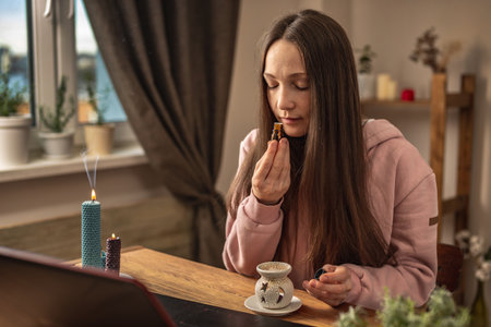 Woman is sitting in front of a laptop, listening to an instructor or a video lecture on meditation and sniffing essential oil from a bottle. Concept of a distance therapy and meditation session.の写真素材