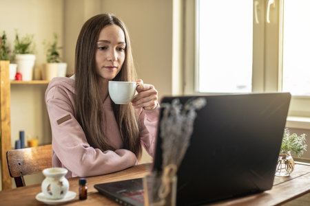 Woman with a cup of tea is sitting in front of a computer screen in a calm pleasant atmosphere and attentively listens to video training or an online session or just communicates with a friend.の写真素材