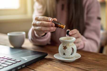Woman is sitting in front of a computer screen and dropping essential oil from a bottle into an aroma lamp to create a pleasant atmosphere and the right mood.の写真素材