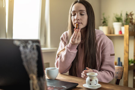 Woman is sitting in front of a computer screen and sniffing essential oil from a bottle to relax and restore energy .の写真素材