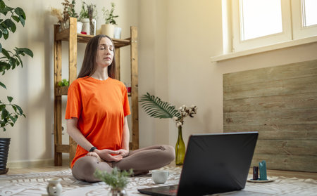 Woman sits in a lotus position on the floor in front of a laptop, meditates and relaxes. Concept of a remote therapy session, meditation and yoga.の写真素材