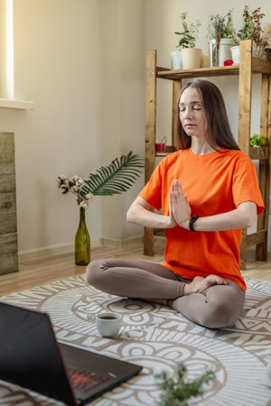 Woman sits in a lotus position on the floor in front of a laptop, meditates and relaxes while listening to an online or a video lecture. Concept of a remote therapy session, meditation and yoga.の写真素材