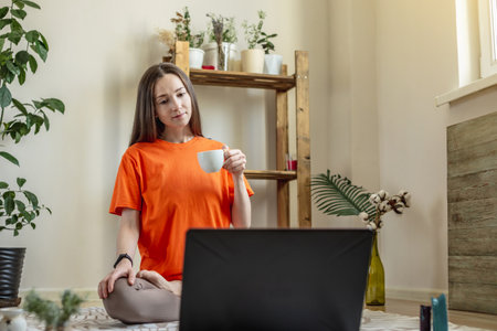 Woman sits on the floor in front of a laptop in the lotus position with a cup of tea in her hands and looks at the screen. Concept of distance or video learning of meditation, yoga.の写真素材