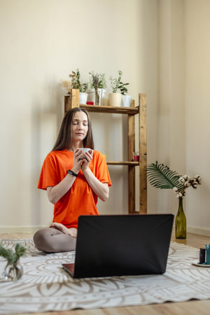 Woman is sitting on the floor in front of a laptop in the lotus position with a cup of tea in her hands with her eyes closed. Concept of a remote therapy and meditation session.の写真素材