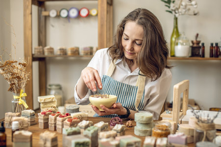 Woman - soap maker in the workshop. A lot of natural soap ready on the table, ingredients and tools on the shelves. A cozy atmosphere of creativity.の写真素材