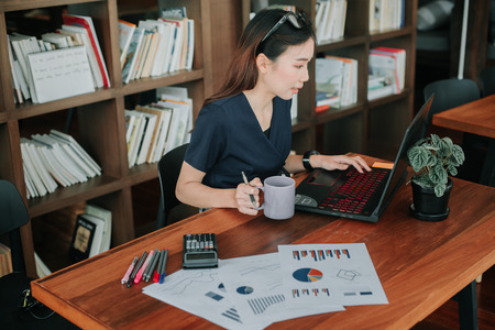 Young Asian businesswoman strictly dressed in the suit working with laptop at the modern cafe...