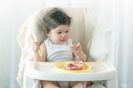 Messy one year baby girl eating cake. Parents leave lovely infant girl holding a spoon, enjoying...
