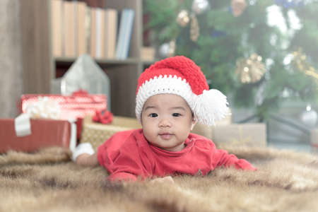 Beautiful little baby celebrates Christmas. New Year's holidays. Baby in red bodysuit and sweater hat laying with blurred christmas tree and gift boxes as background.の写真素材