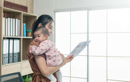 Busy woman trying to work while babysitting newborn baby daughter.  Young beautiful Asian mother holding little baby on shoulder while another holding and watching paper another hand.の写真素材