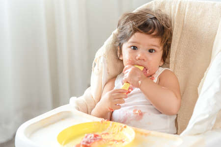 Messy baby girl eating cake.  Lovely infant girl holding a spoon, enjoying eating cake.