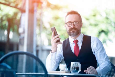 Middle age businessman sitting at a cafe drinking coffee and using smart phone. Caucasian handsome man holding mobile phone smiling to camera at coffee shop.の写真素材