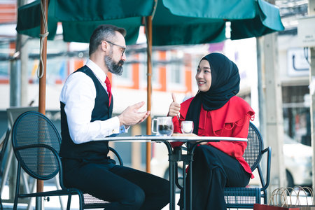 Handsome beard middle age caucasian businessman drinking coffee at outdoor coffee cafe with young beautiful hijab muslim women. Focus on women smiling.の写真素材
