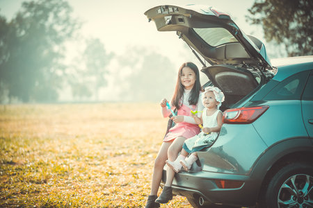 Girl and her infant sister sitting on back side of car playing with soap bubbles in park.の写真素材