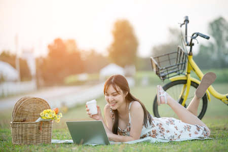 Beautiful young Asian woman having picnic on sunny spring day in park, happy reading things on internet, having cup of coffee, bicycle blurred background.の写真素材