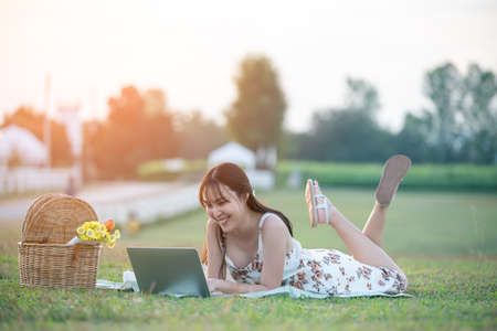 Beautiful young Asian woman having picnic on sunny spring day in park, happy reading things on internet, having cup of coffee.の写真素材