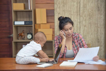 Pretty young single mom working at home, talking with customer on the phone and holding the box while little daughter playing a laptop on desk.の写真素材