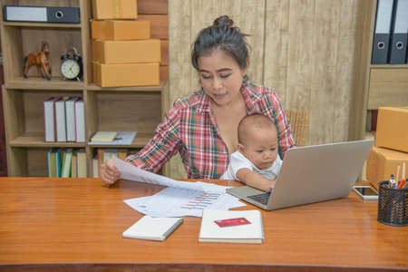 Pretty young single mom working at home on a laptop computer while holding her baby girl sitting on her lap enjoying watching mother working.の写真素材