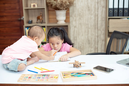 Little toddler girl sit on the working table, her older sister sitting and playing together while mother not homeの写真素材