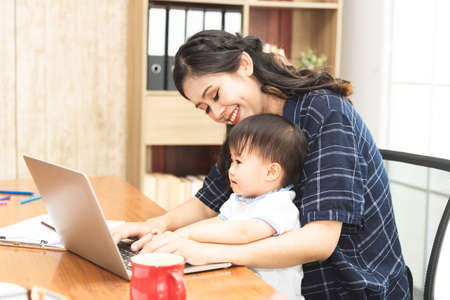 Beautiful Asian business mom is using a laptop  while spending time with her cute baby boy at home for quarantine from virus crisis, work from home concept.の写真素材
