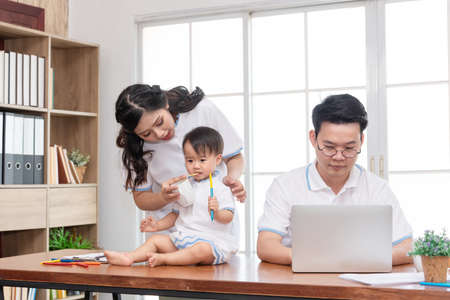Asian mother taking care of baby while father working on computer, work from home family. Mother feeding baby.の写真素材
