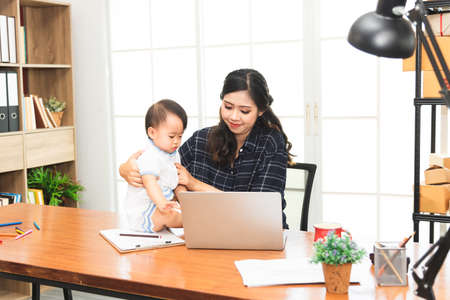 Beautiful Asian business mom is using a laptop  while spending time with her cute baby boy at home for quarantine from virus crisis, work from home concept.の写真素材