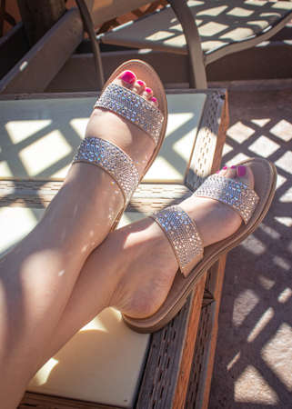 Feet with a pedicure and bedazzled shoes resting crossed on outdoor furniture with lattice shadowsの写真素材