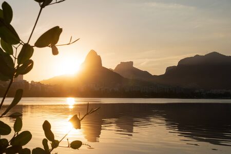 Golden sun dips below the horizon against Morro Dois Irmaos Two Brothers Mountain and Pedra da Gavea, reflected in Lagoa Rodrigo de Freitas Lake, Rio de Janeiro, framed by silhouetted leaves and sky.の写真素材