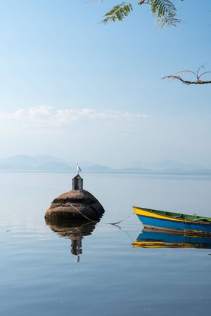 A small colored timber boat is tied to a rock and reflected in calm waters of Ilha de Paqueta Island in Rio de Janeiro. There is a white bird on the rock, wth blue sky and mirror like sea.の写真素材