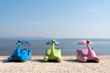 A row of colored swan paddle boats on the sand at Ilha de Paqueta Island in Rio de Janeiro. The beach has blue calm water with mountains beyond on a sunny afternoon. The boats are facing the shore.の写真素材