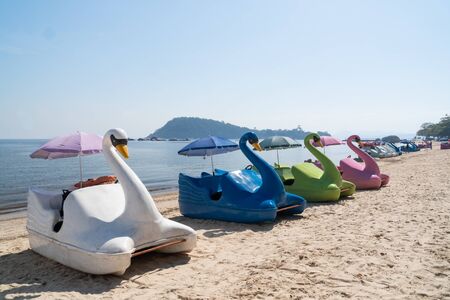 A row of colored swan paddle boats on the sand at Ilha de Paqueta Island in Rio de Janeiro. The beach has blue calm water with mountains beyond on a sunny afternoon. The boats are facing the shore.の写真素材