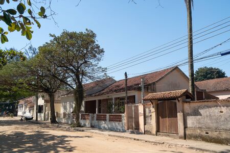 A quiet dirt road on the Ilha de Paqueta Island in Rio de Janeiro. The residential street has old colonial houses with tile roofs, front walls, gates and trees on a sunny afternoon with blue sky.の写真素材