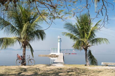 A white clock tower lighthouse at the end of a narrow concrete pier on Ilha de Paqueta Island, Rio de Janeiro. There are palm trees either side, with calm blue sea, small fishing boats and a bike.の写真素材