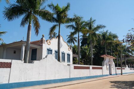 Rio de Janeiro, Brazil - July 25, 2019: A large colonial house on a dirt road on Ilha de Paqueta Island. The building is white with blue shutters and a tile roof, with palm trees and a tall fence.のeditorial素材