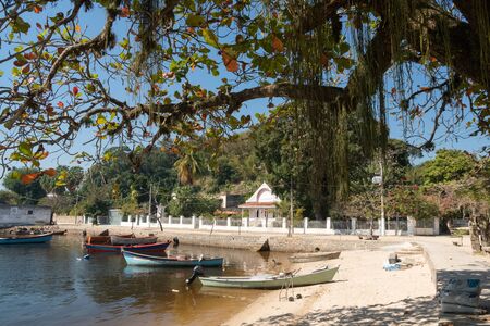The waterfront on Ilha de Paqueta Island, Rio de Janeiro, There is calm brown water, a small white sand beach, overhanging tree, small colored fishing boats, blue sky and white buildings on a street.の写真素材