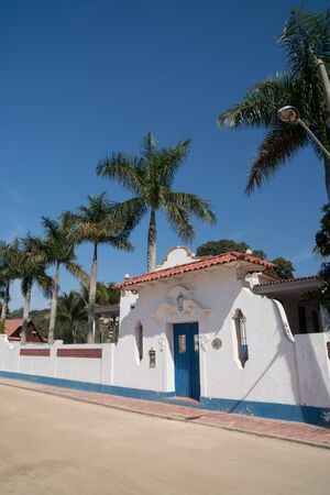 Rio de Janeiro, Brazil - July 25, 2019: A large colonial house on a dirt road on Ilha de Paqueta Island. The building is white with blue shutters and a tile roof, with palm trees and a tall fence.のeditorial素材
