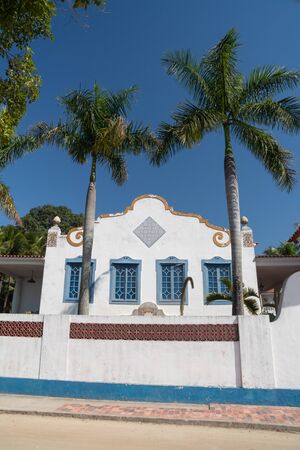 Rio de Janeiro, Brazil - July 25, 2019: A large colonial house on a dirt road on Ilha de Paqueta Island. The building is white with blue shutters and a tile roof, with palm trees and a tall fence.のeditorial素材