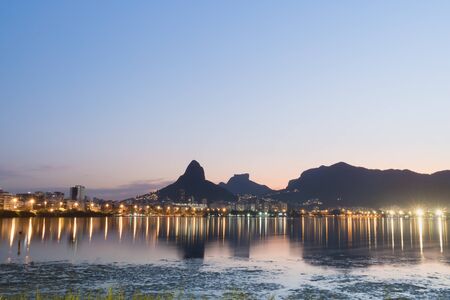 Sunset from the lakeside edge of Lagoa Rodrigo de Freitas in Rio de Janeiro, with clear blue and pink sky and street lights. Dois Irmaos Mountain and Pedra da Gavea are reflected in the lake water.の写真素材