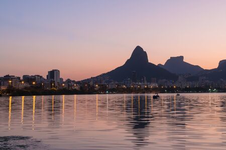 Sunset from the lakeside edge of Lagoa Rodrigo de Freitas in Rio de Janeiro, with clear pink sky and orange street lights. Dois Irmaos Mountain and Pedra da Gavea are reflected in the calm lake water.の写真素材