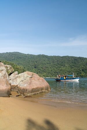 Early morning at Praia do Pouso Beach, Ilha Grande island in the state of Rio de Janeiro, Brazil, on the trail to Praia Lopes Mendes. There is golden sand, rocks, a boat, green mountains and blue sky.の写真素材