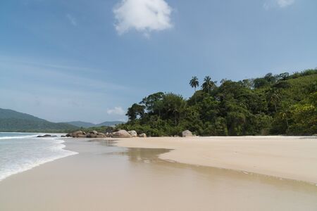 Early morning at the deserted Praia Lopes Mendes on Ilha Grande Island in the state of Rio de Janeiro, Brazil. There is blue sky, white sand, small waves, palm trees, mountains beyond, with no people.の写真素材