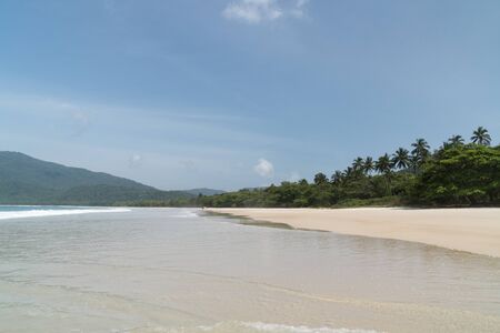Early morning at the deserted Praia Lopes Mendes on Ilha Grande Island in the state of Rio de Janeiro, Brazil. There is blue sky, white sand, small waves, palm trees, mountains beyond, with no people.の写真素材