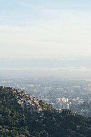 The Morro dos Prazeres favela in Rio de Janeiro from Mirante Dona Marta in the early morning. The sunrise light shines on the low fog, with the city beyond. The slum is very poor with brick houses.の写真素材