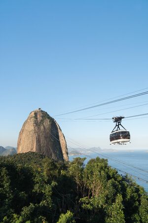 A Sugarloaf Mountain cable car in the air over tropical trees in Rio de Janeiro on a sunny evening. There is blue sky and sea, with light shining on the steep granite peak.の写真素材