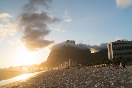 Rio de Janeiro, Brazil - January 2, 2020: A golden sunset from Sao Conrado Beach. Pedra da Gavea, a tall granite rock mountain sits at the end of the urban beach, and people relax in the white sand.のeditorial素材