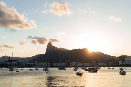 Golden sun peaking over the edge of the Tijuca Forest Mountains, Corcovado and Christ the Redeemer Statue in Rio de Janeiro, seen across Guanabara Bay from Mureta da Urca. There is blue sky and boatsの写真素材