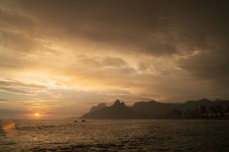 A dramatic yellow and orange moody sunset over Ipanema Beach and Morro Dois Irmaos Two Brothers Mountain in Rio de Janeiro. There is a low sun with golden light shining up onto dramatic stormy cloudsの写真素材