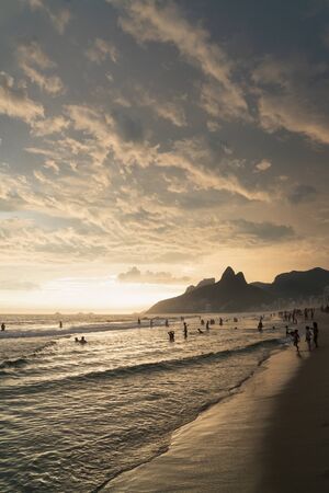 A dramatic sunset over Ipanema Beach in Rio de Janeiro, Brazil,with golden light from the low angle sun shining up onto the stripes of grey clouds. There are silhouettes of people swimming in the sea.の写真素材